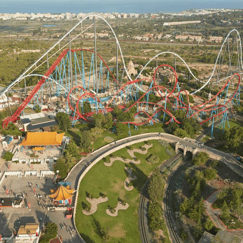 Immagine che rappresenta il parco PortAventura visto dall'alto, con il particolare di due montagne russe una rossa (Shambhala) e una bianca, dei giardini e il mare sullo sfondo. Image showing PortAventura park seen from above, with a detail of two roller coasters, one red (Shambhala) and one white, some gardens and the sea in the background.