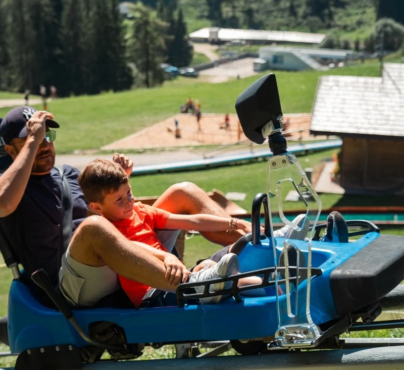 Immagine che ritrae un adulto e un bambino mentre sono sopra ad un bob su rotaia all'Alpine Coaster Gardoné a Latemar di Predazzo. Image showing an adult and a child while they are on a bobsleigh on rails at the Alpine Coaster Gardoné in Latemar di Predazzo.