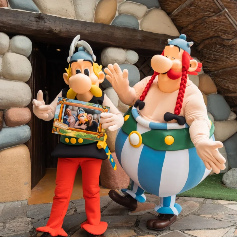 Immagine che ritrae Asterix e Obelix in un parco divertimenti che tengono in mano una foto stampata. Image showing Asterix and Obelix in an amusement park holding a printed photo.